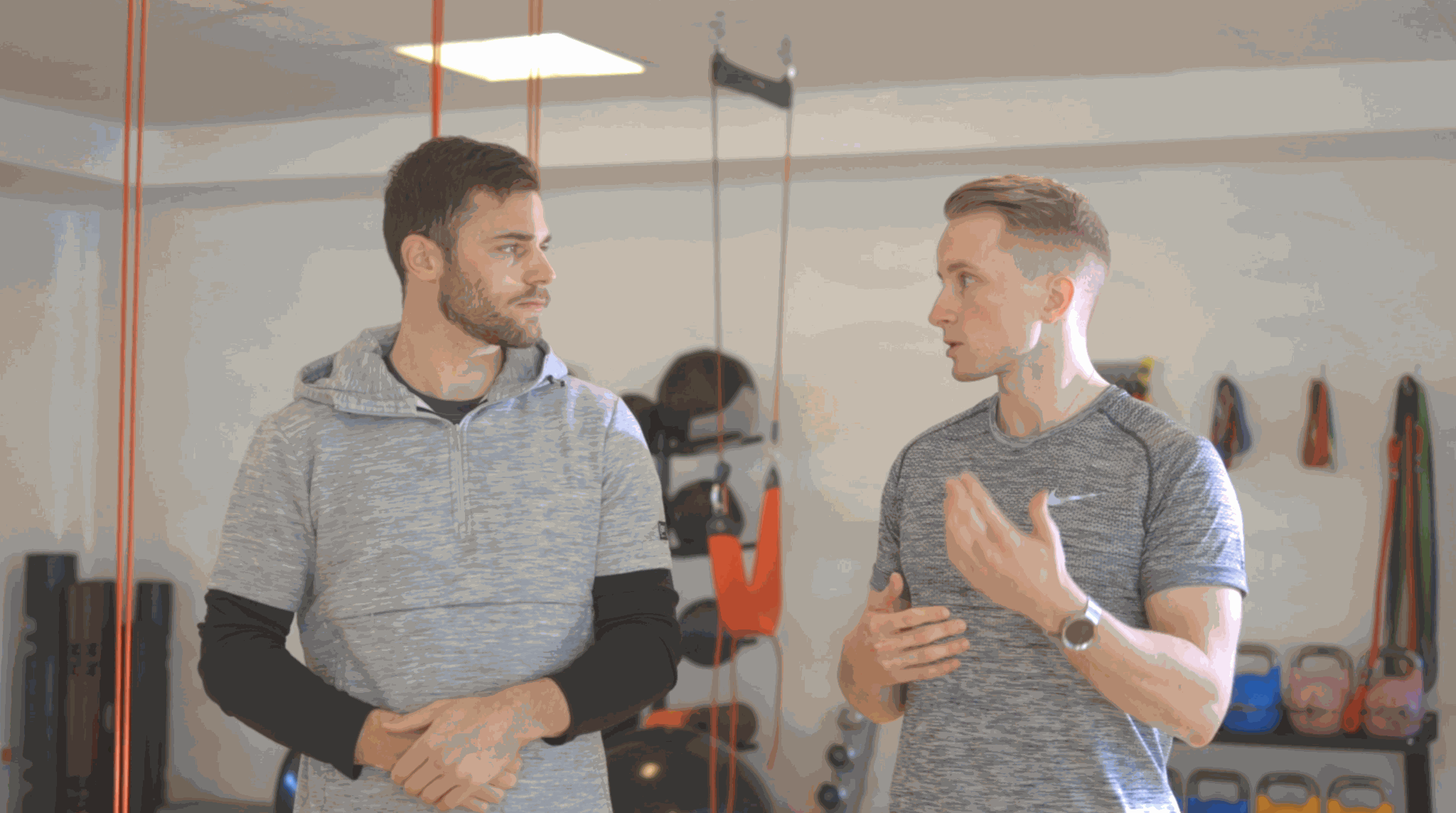 Two men having a conversation in front of an A90 Prehab: Self-Screening & Corrective Exercises (Video Course) in a gym equipped with various fitness gear.