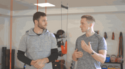 Two men having a conversation in front of an A90 Prehab: Self-Screening & Corrective Exercises (Video Course) in a gym equipped with various fitness gear.